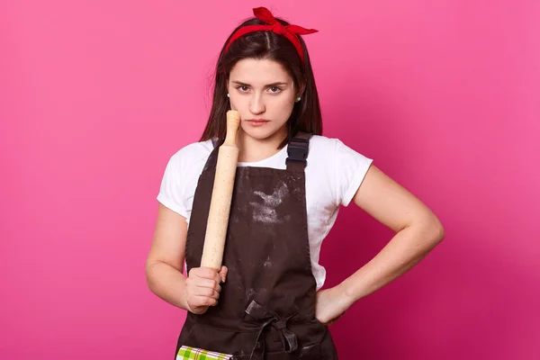 La hembra seria y delgada sostiene el rodillo para hornear, usa delantal marrón untado con harina, camiseta blanca y diadema roja. Joven linda chica tiene mirada dura cansado. Cocinero aislado sobre fondo rosa . — Foto de Stock