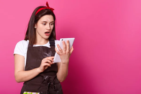 Wanita yound terguncang berdiri dan menonton berita pada tablet putih, terlihat sangat terkejut menonton dengan mulut terbuka secara luas. Sincere perempuan memakai pakaian kerja, terlihat berantakan dengan tepung . — Stok Foto