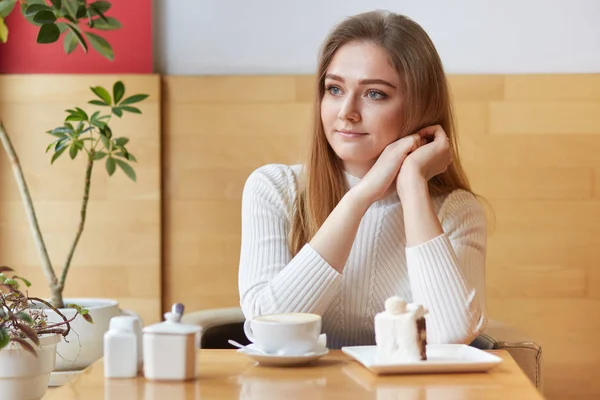 Modelo bonito senta-se no café local, olha na janela, come delicioso bolo e bebe café, coloca a cabeça nas mãos, se sente satisfeita. Jovem terno tendo descanso em ambiente acolhedor e agradável . — Fotografia de Stock