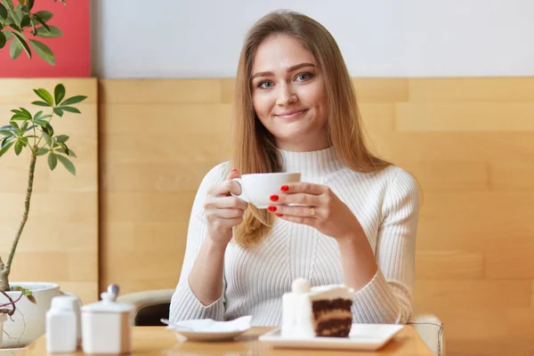 Jovem encantadora aquece no café bebendo chá quente, segura xícara nas mãos, vai comer bolo saboroso, usa vestido branco inteligente. Mulher bonita parece satisfeito passar minuto livre neste lugar . — Fotografia de Stock