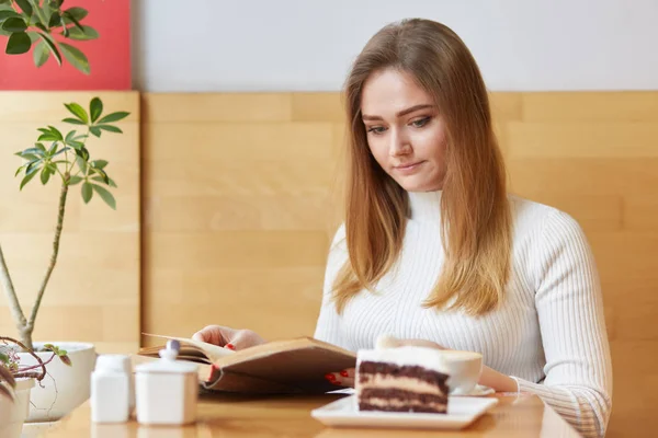 Um retrato da menina atraente séria senta-se no café local e lê livro velho, veste roupas brancas. Jovem de cabelos bonitos passa seu tempo de lazer no café com xícara de café e bolo delicioso . — Fotografia de Stock