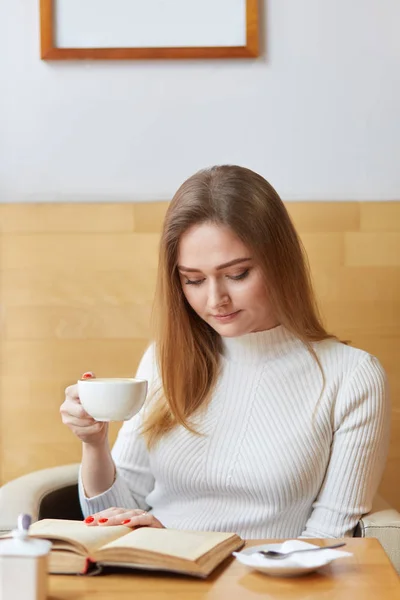 Young model poses sitting in cafe, reading interesting book, holding cup of coffee in right hand, focusing on details of plot. Good looking slender girl feels comfortable and spends her free time. — Stock Photo, Image