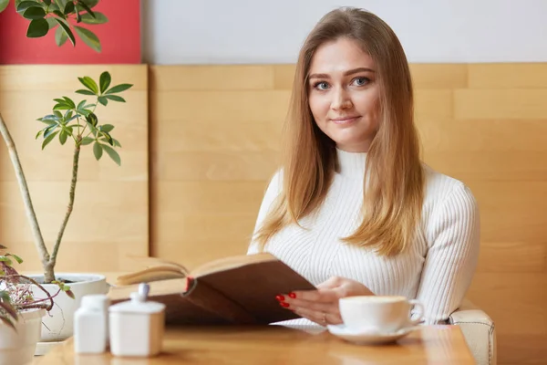 Adorável visitante loiro do café local usa vestido branco, come bolo saboroso cremoso, bebe xícara de café. Sincero modelo bonito posa com livro velho marrom em suas mãos. Conceito de atividades de tempo livre . — Fotografia de Stock