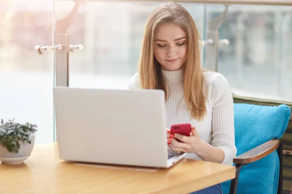 Concentrada menina estudante loira de cabelos longos verificando a rede social com telefone enquanto descansa no café. Jovem senhora senta-se à mesa no café e trabalha online, usa wi-fi gratuito. Pessoas e tecnologia . — Fotografia de Stock