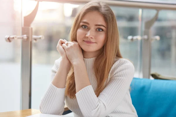 Portrait of attractive young girl being in good mood, has rest in cafeteria after lectures in university, sits on comfortable blue sofa, wears casually, looks at camera, keeps hands folded under chin. — Stok fotoğraf