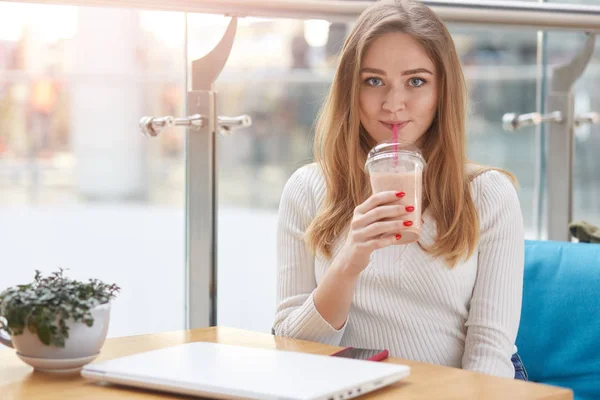 Encantadora jovem loira bebendo coquetel enquanto descansa depois do trabalho no livro líquido, sentada no café, freelancer mulher fazendo uma pausa após o site de desenvolvimento, usando Internet sem fio, olha para a câmera . — Fotografia de Stock