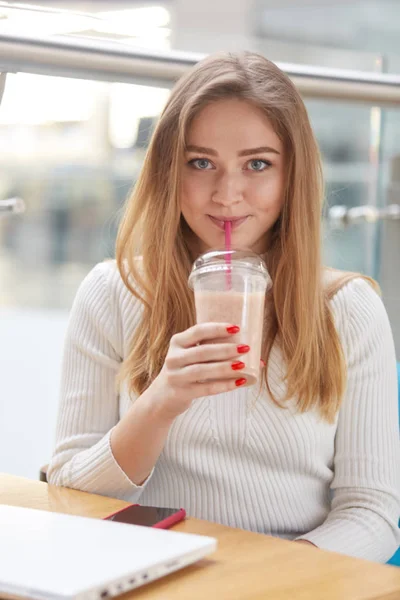 El retrato vertical de la hermosa chica youn se viste casualmente, se sienta en un restaurante o cafetería, bebe cóctel mientras descansa después de trabajar en línea, tiene una expresión facial agradable. Concepto juvenil . — Foto de Stock