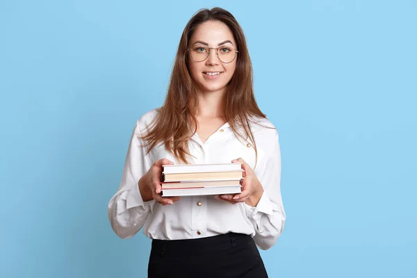 Charismatic cheerful girl holds bunch of books in both hands, wears formal in white blouse and black skirt, fashionable spectacles. Cute attractive lady is in high spirits to study and practice. — Stock Photo, Image