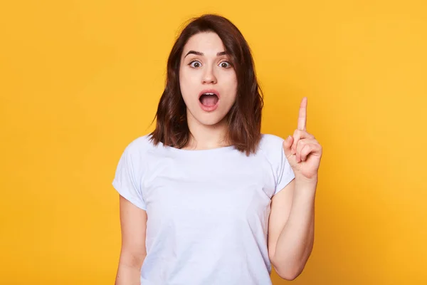 Boa aparência feminina morena vestida casual t-shirt, olha em espanto como indica algo para cima, isolado sobre fundo amarelo. Jovem encantadora vê uma coisa incrível. Espaço de cópia . — Fotografia de Stock