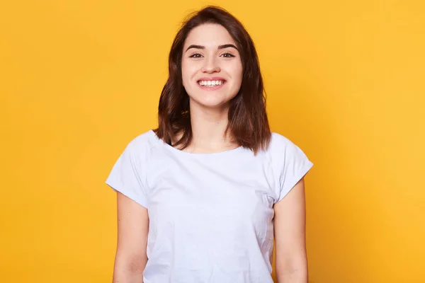 Retrato de mujer caucásica atractiva emotiva se ríe mientras mira directamente a la cámara y de pie sobre el fondo amarillo. Encantadora morena hembra con sonrisa dentada se ve feliz, tiene grandes noticias . — Foto de Stock