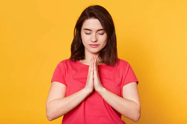 Belle jeune femme brune fait un geste de prière, garde les paumes serrées, se sent coupable, vêtue d'un t-shirt rouge décontracté, demande pardon, isolée sur fond jaune. Concept de personnes . — Photo