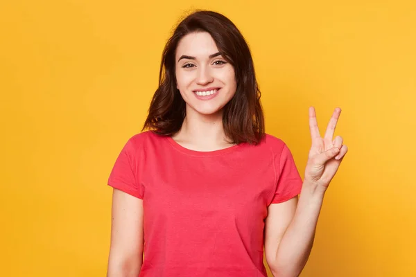 Sorrindo bela jovem vestida casualmente, mostrando dois dedos ou gesto de vitória, isolado sobre fundo amarelo, tem expressão facial feliz, parece satisfeito, posa com sorriso de dente . — Fotografia de Stock