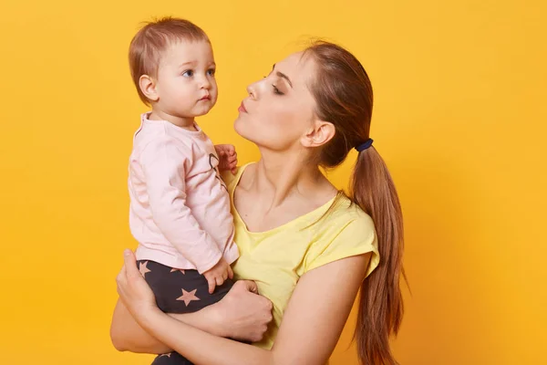A young caring mother tries to calm down her crying daughter. A tear is going to run down cute baby`s cheek. A disappointed child looks concentrated on something aside. Mum kisses her amazing kid. — Stock Photo, Image