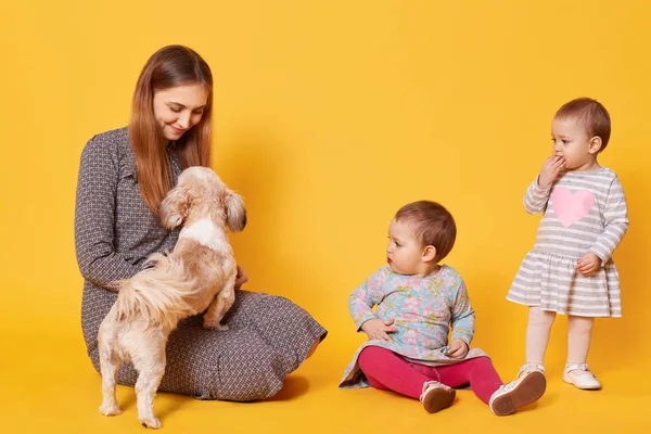 Una madre feliz y encantada juega con su mascota favorita, sentada en el suelo, mirándola. Las hijas gemelas sorprendidas tienen miedo de ir hacia el perro y tocarlo. Concepto de tiempo libre familiar . — Foto de Stock