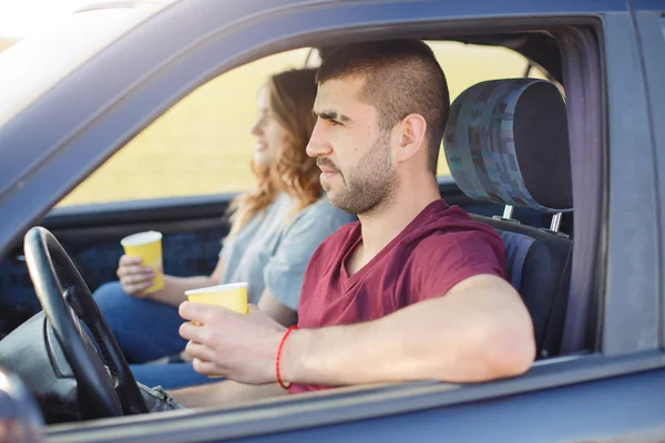 Young couple with coffee inside of car, have rest riding along road, happy family spend time together, travel on weekend, have trip or journey, side view of male and famale being in traffic jam.