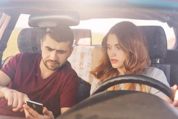 Puzzled couple sits in automobile, bearded man holds smart phone, uses online maps, tries to find way, being lost, have journey on weekend. Family in automobile chek email while parking on road side.