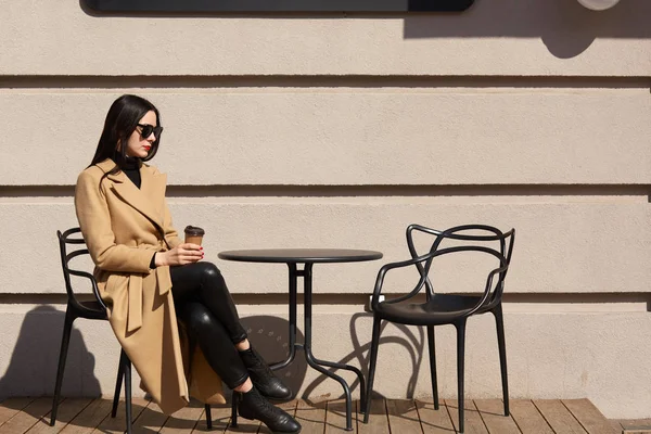 Ta bort kaffet i kvinnors händer. Vacker ung brunett kvinna klädd i brun kappa sitter vid bordet i gatan utomhus kafé och dricka varm dryck, åtnjuter soliga höst dagar, ser thougtful. — Stockfoto