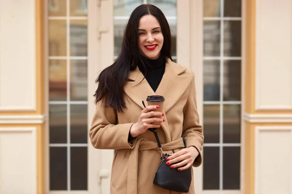 Joven hermosa morena bebe café mientras camina por la ciudad, vestido elegante abrigo beige, pequeño bolso, tiene labios de color rojo brillante. Posando contra la ventana del edificio. Concepto de personas . — Foto de Stock