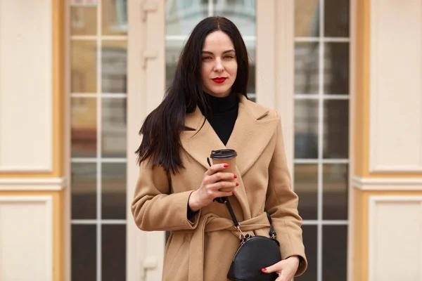 Retrato de mujer elegante atractiva con abrigo beige y beber café para llevar mientras camina por la calle de la ciudad. Joven morena lokks directamente en la cámara, tiene misteriosa expresión facial . — Foto de Stock