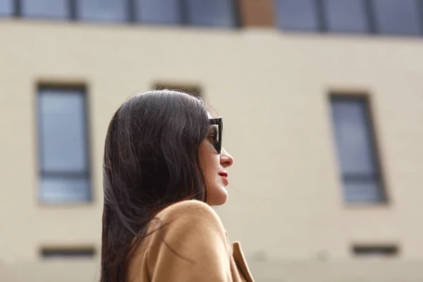 Портрет красивой молодой предпринимательницы на открытом воздухе на фоне размытой улицы. Профиль элегантной брюнетки женского пола, носит бежевое пальто и черные солнцезащитные очки, смотрит прямо вперед. Концепция . — стоковое фото