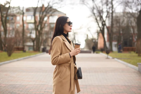 Mulher morena na moda passa o dia de folga andando na cidade. Fofa encantadora fêmea segura uma xícara de papel de bebida quente em sua mão, impressionado pela vista urbana. Conceito de moda, beleza e estilo . — Fotografia de Stock