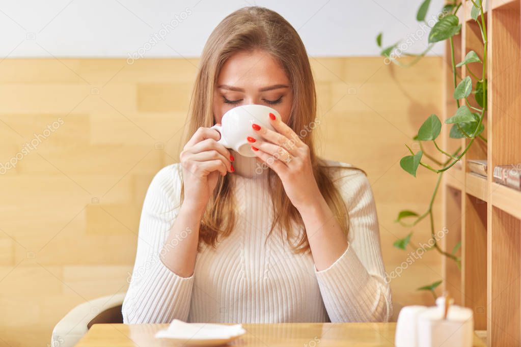 Portrait of positive calm female sipping hot drink in cafe with wooden stylish interior, holds cup in hands tightly, spends weekends in warm convenient place. Free time activities concept.
