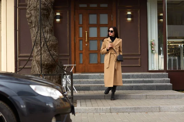 Foto de cuerpo entero de una hermosa morena de pie cerca del edificio de la ciudad, bebiendo café caliente para llevar, yendo a su coche, mira a un lado, usando abrigo beige, botas, gafas de sol, pasa tiempo libre . — Foto de Stock