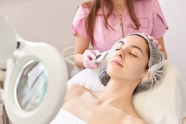 Beautiful woman receiving ultrasound facial peeling. Skin cleansing procedure at beauty spa salon. Cosmetologist makes cleansing treatment for her young client via moder equipment. Skin care concept.