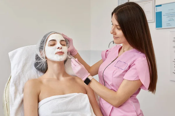 Wizerunek młodej kobiety z maską peeling twarzy, zabiegi kosmetyczne spa. Kobieta coraz pielęgnacji twarzy przez kosmetyczki w salonie spa. Kosmetolog stosuje środek kosmetyczny ze specjalną szczotką. Koncepcja pielęgnacji skóry. — Zdjęcie stockowe