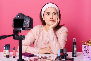 Çekici düşünceli Kafkas vlogger yakın portresi, kozmetik ile masada tripod üzerinde kamera ile video filme, online translilasyon vardır, viwer bekler, düşünceli yüz ifadesi vardır.