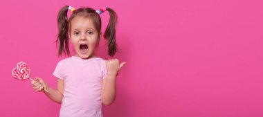 Pigtails ile duygusal çığlık küçük kız başparmak ile yön gösterir, geniş ağzını açar, terör ile gözleri açar, diğer elinde lezzetli tatlı lolipop tutarak. Çocukluk ve aktivite kavramı.