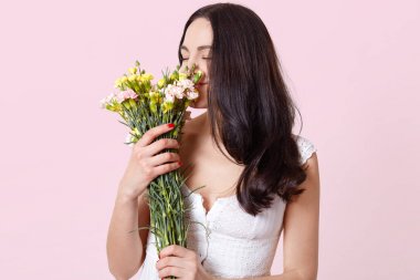 İhale tatlı esmer genç bayan düz ayakta, her iki elinde güzel çiçekler tutarak, onların bahar kokusu hissi, memnun görünüyor, beyaz yazlık elbise giyiyor. İnsanlar ve duygular kavramı.