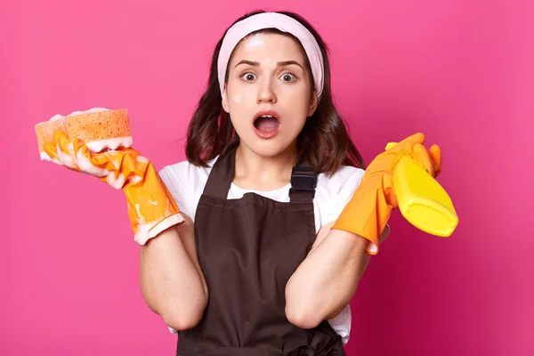 Studio shot of surprised female housekeeper holds sponge and detergent spray in hands, having much work to do, uses cleanser, looks positively, wearing brown apron and orange protective gloves. — Stock Photo, Image