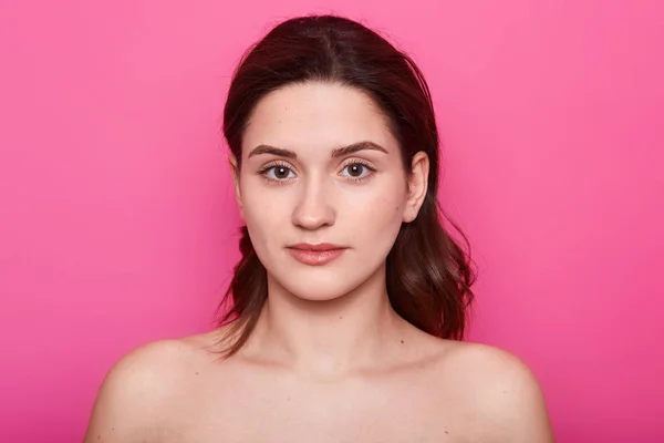 Close up portrait of beautiful young caucasian brunette woman with clean skin after procedures in spa salon, shows bare shoulders, looks calm, posing aginst pink wall. Beauty and skin care concept. — Stock Photo, Image