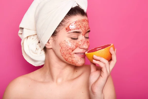 Prise de vue en studio d'une jeune femme européenne agréable à regarder sentant le pamplemousse, a une serviette blanche sur la tête, montrant ses épaules nues, posant isolée sur fond rose. Concept beauté et soin de la peau . — Photo