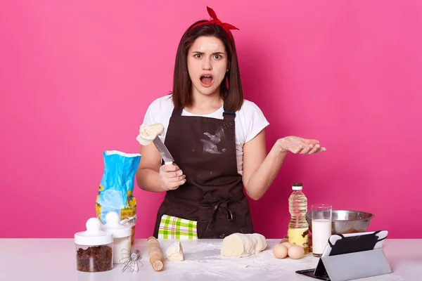 Estudio de toma de mujer sorprendida panadero corte de masa en la cocina casera, sostiene cuchillo con parte de la masa, se para cerca de la mesa con diferentes productos, la preparación para las vacaciones de Pascua, aislado sobre fondo rosa — Foto de Stock