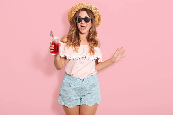 美丽的年轻女子在夏天上衣，短，帽子和太阳镜，拿着壶与冷饮，而站在粉红色的背景，恳求花她的假期在海边与她的朋友. — 图库照片