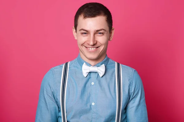 Laughing hilarious man stands straight in front of camera, gives cunning cheery expression. Sprightly jolly young man wears blue shirt, white bow tie, spenders. Copy space for advertisement. — Stock Photo, Image