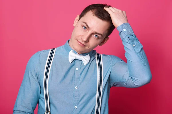 Charismatic handsome young man touches his hair with hand, tilts his head in one side, isolated over pink studio background. Blue eyed athletic man wears blue shirt, white bow tie, spenders.