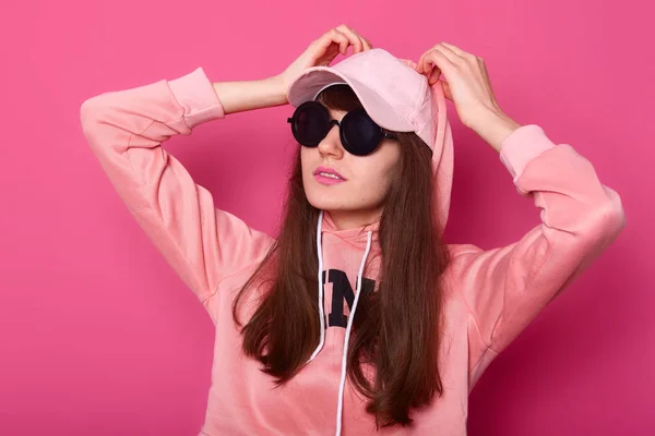 Jonge mode model tienermeisje poseren in de studio in stijlvolle roze hoody, pet en balck zonnebril, houdt handen op de motorkap. Casual stijl, schoonheid accessoires. High fashion casual stijl, roze achtergrond. — Stockfoto