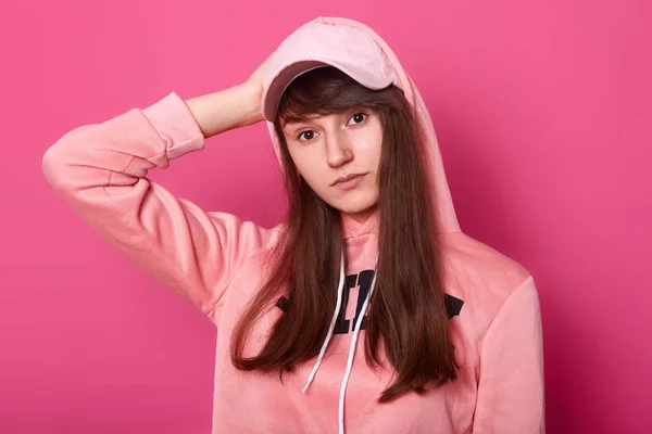 Beeld van brunette meisje kijkt naar de camera met ernstige en thoufgtfull gezichtsuitdrukking, het dragen van casual roze Hoody en Cap geïsoleerd op Rose achtergrond, tiener poses in fotostudio. Modellering concept. — Stockfoto