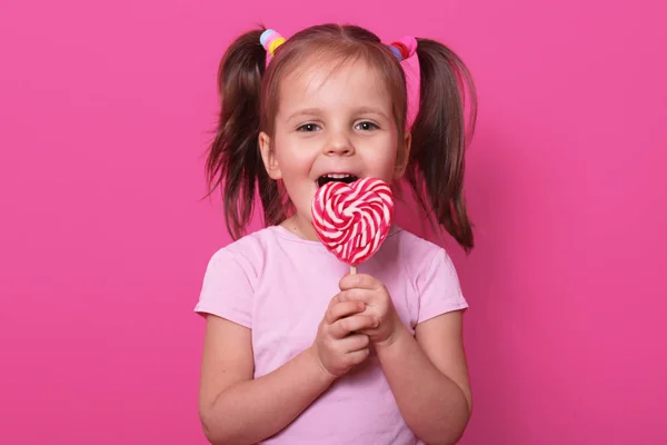 Gelukkig schattig meisje draagt Rose t Hirt, staat geïsoleerd over roze achtergrond, houdt heldere Lollipop in handen. Vrolijk kind met geopende mond proeverij heerlijke snoep. Jeugd en smaken concept. — Stockfoto