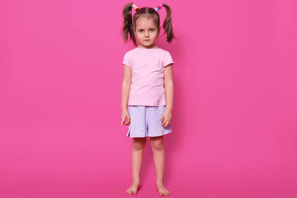 La pequeña chica magnética cansada con coletas se centra en la cámara, se ve seria, con una camiseta de color rosa claro y pantalones cortos casuales, que tiene scrunchies coloridos en su cabello. Concepto de infancia y emociones . —  Fotos de Stock