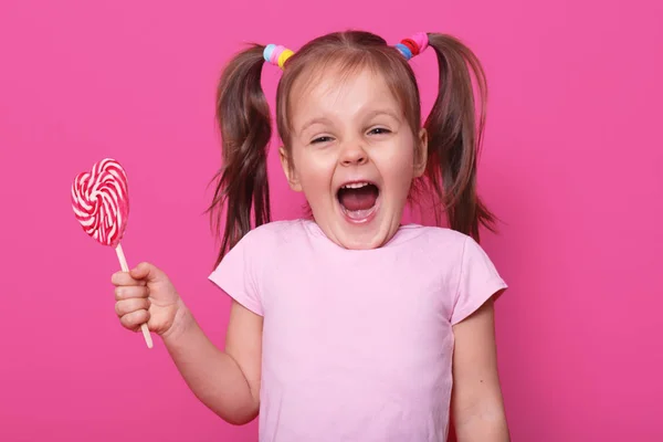 Laughing screaming cute girl opens her mouth widely, showing teeth, holds in one hand heart bright tasty lollipop. Emotional positive childen spends her leisure time with pleasure. Copy space for ad. — Stock Photo, Image