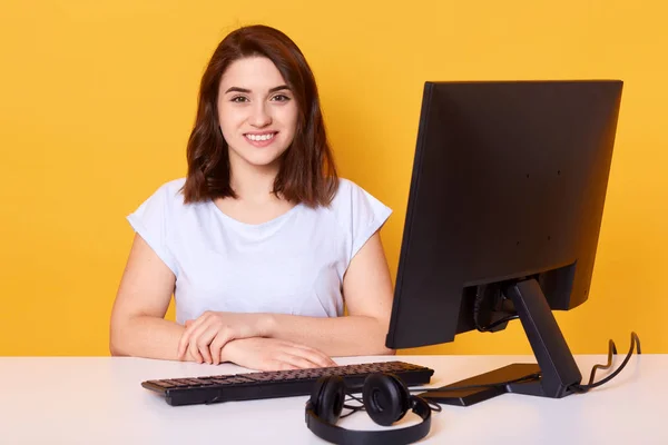 コンピュータで働く幸せな陽気な若いブルネットの女の子の肖像画は、インターネットでお金を稼いで、カジュアルに服を着て、カメラを直接見て、黄色の背景の上に孤立したポーズ。技術コンセプト. — ストック写真