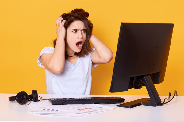 Surprised beautiful brunette girl with hands on head and opemouth sitting in front of computer and looking at screen with shocked expression reading some unexpected news on internet, wearing casually.