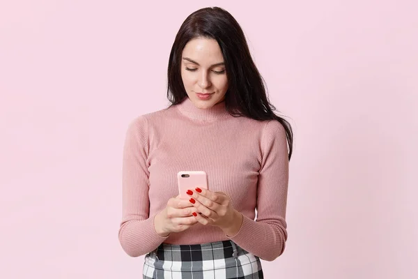 Dulce atractiva joven sonriente se encuentra aislada sobre fondo rosa, mirando su teléfono móvil, leyendo las últimas noticias, vistiendo jersey rosa, falda en blanco y negro. Copiar espacio para publicidad . — Foto de Stock