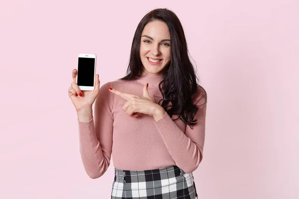 Модная веселая молодая женщина держит новый мобильный телефон, направляет указательный палец на него, презентуя покупку, выглядит счастливой. Восхитительная милая улыбающаяся модель позирует изолированно на светло-розовом фоне . — стоковое фото