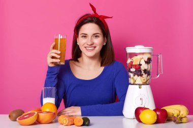 Gülümseyen samimi kadın mavi sweatshirt ve kırmızı kafa bandı giyen, onun sağlığı için besleyici yararlı karışımı ile cam tutan, olumlu ve enerjik görünüyor, pembe parlak arka plan üzerinde izole.