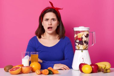 Şaşkın şok kız şüpheli bir görünüm ve yaygın olarak açılan ağız ile kameraya doğrudan bakar, kırmızı trendy kafa bandı ve koyu mavi kazak giyiyor, gerçekler ve diyet planı ile şok. Yaşam tarzı kavramı.